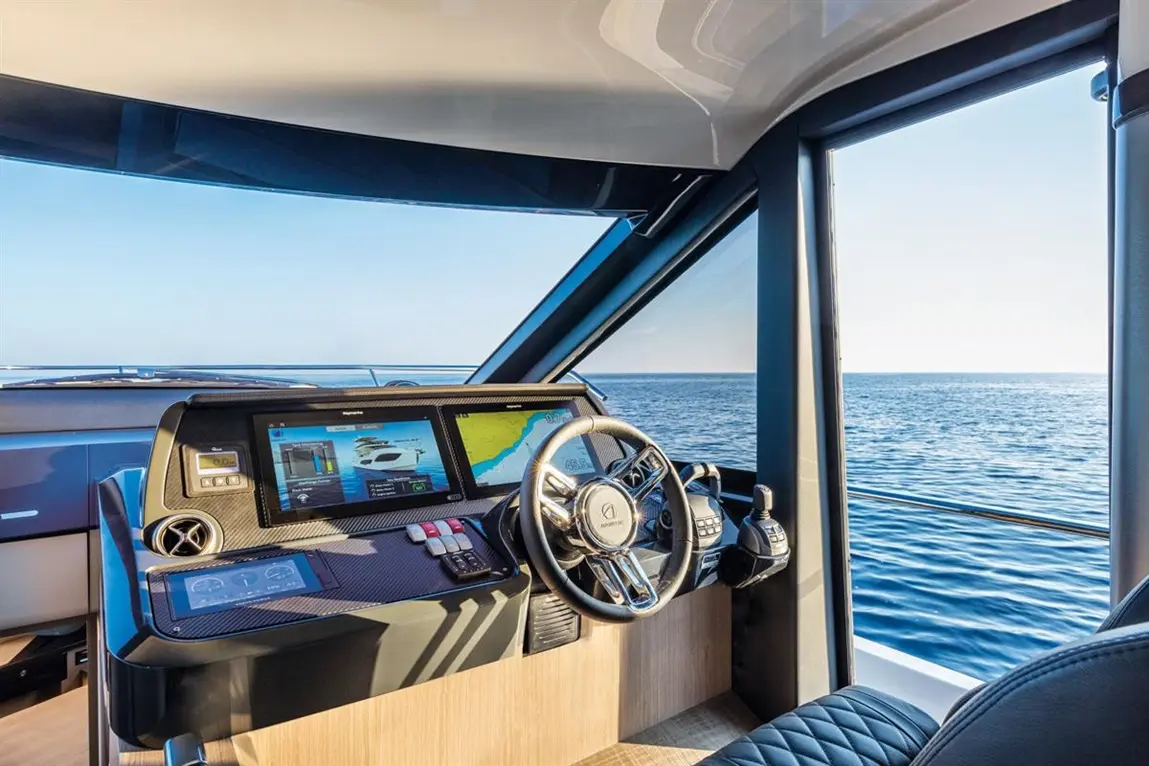 Modern yacht cockpit featuring two digital screens, a steering wheel, and a clear view of the calm sea.